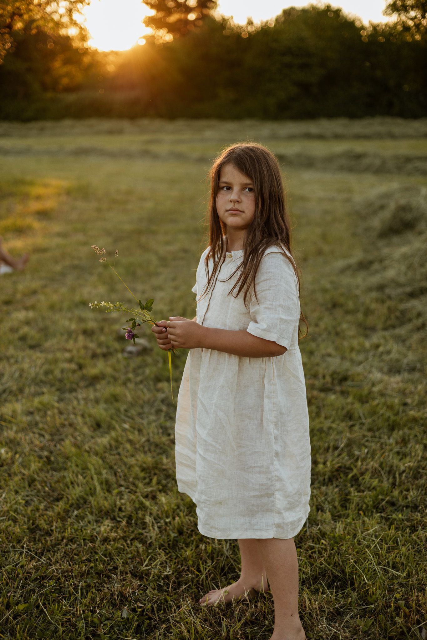 Young girl in white linen dress holds wildflowers barefoot in a sunlit meadow at golden hour.