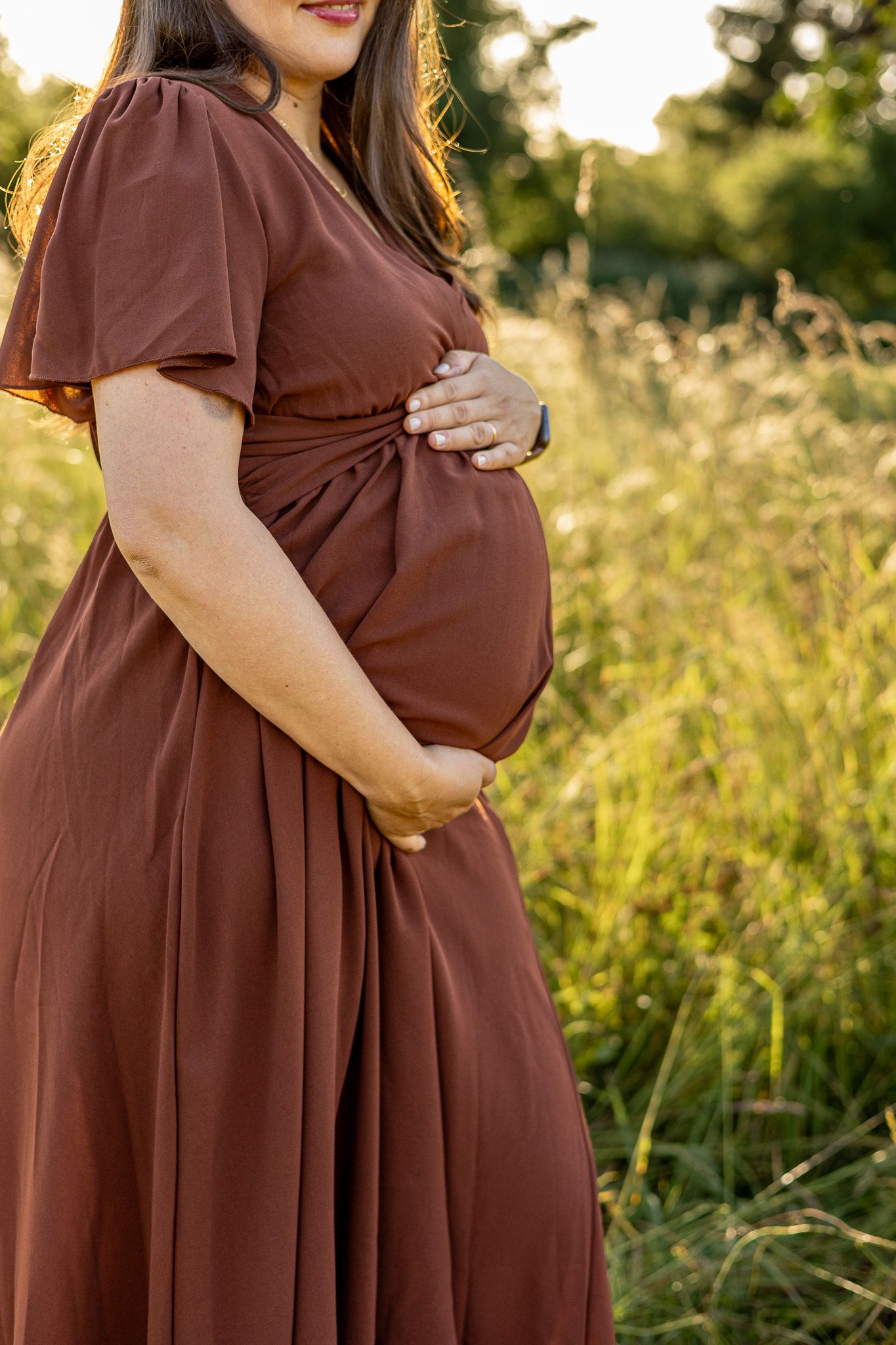 Pregnant woman in brown dress cradling baby bump in sunlit field.