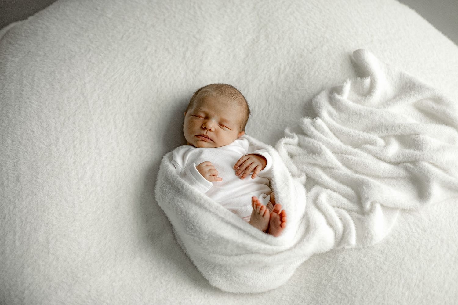 Peaceful newborn baby swaddled in white blanket sleeping on soft white surface.