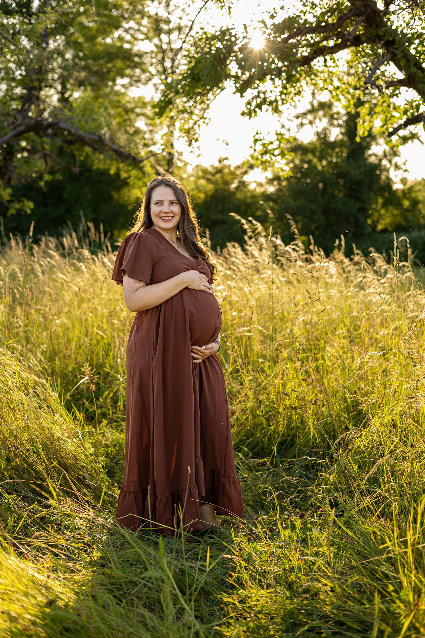 Smiling pregnant woman in brown maxi dress cradling baby bump in golden sunlit meadow with lush green trees.