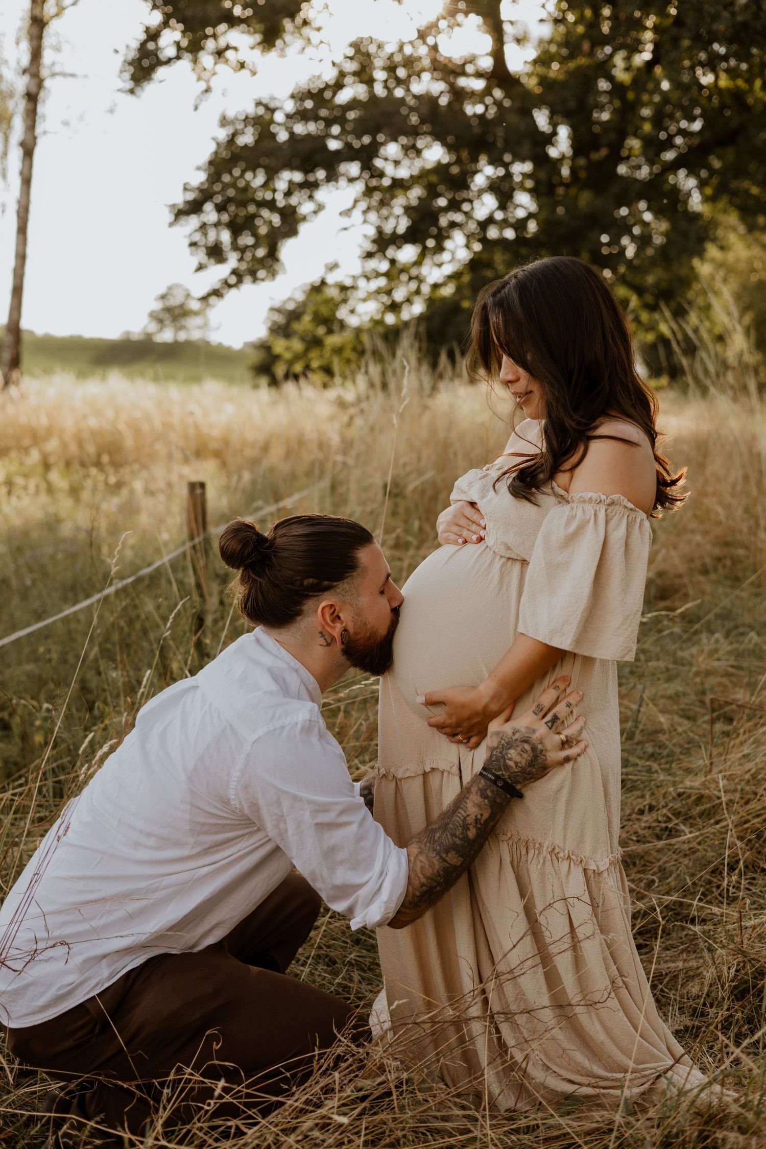 A tattooed man kneels kissing a pregnant woman's belly in a golden field during a maternity photoshoot.