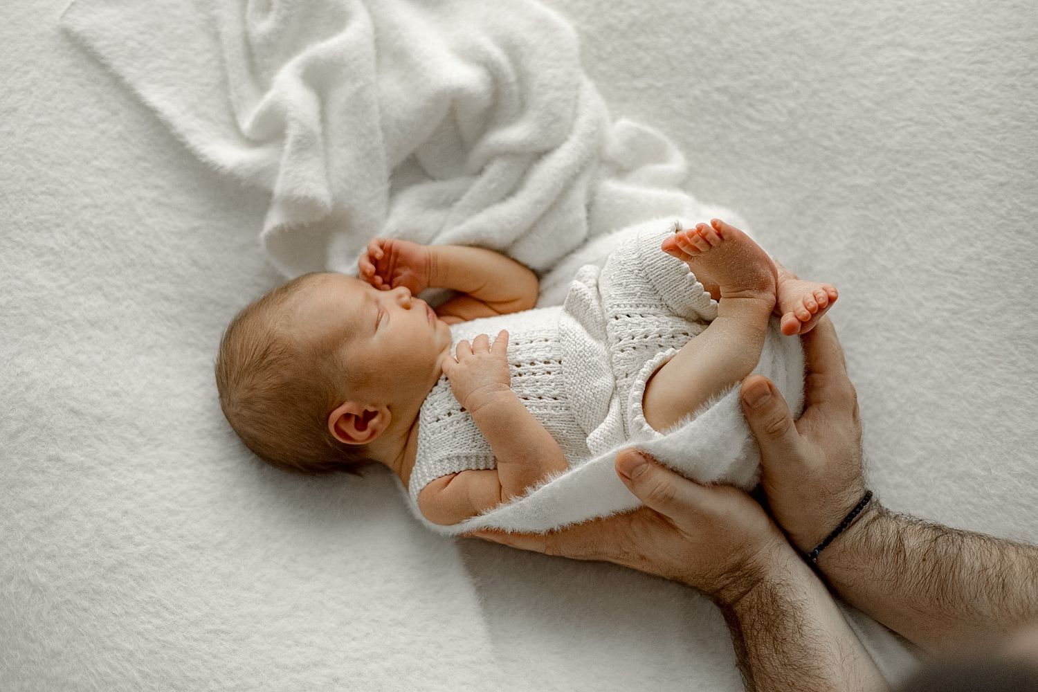 Newborn baby in white knit outfit lying on soft blanket while adult hands gently cradle the infant.