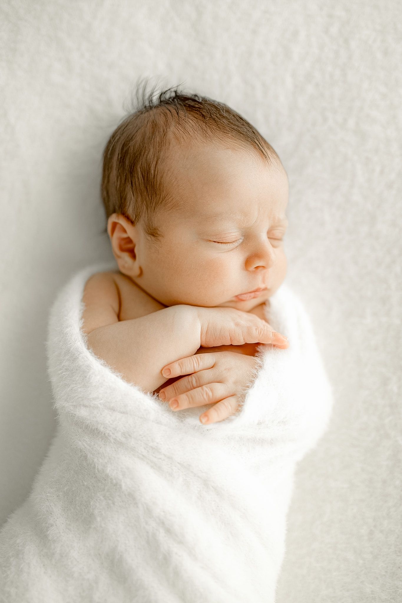 Peaceful newborn baby sleeping swaddled in a soft white blanket with tiny hands folded under chin.