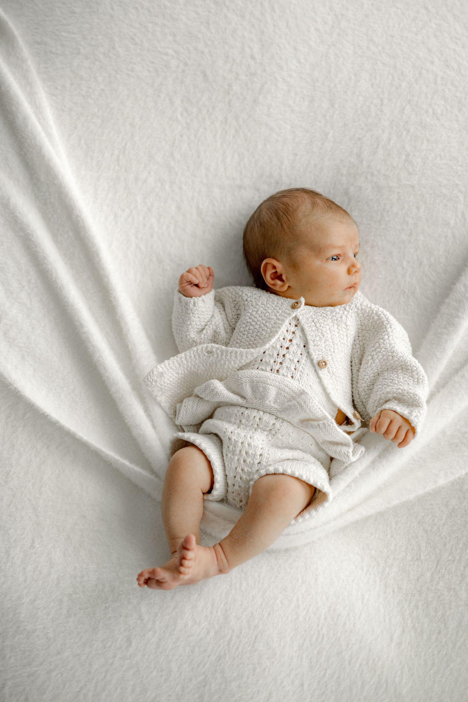 Newborn baby in white knit cardigan and shorts lying on soft white blanket, peaceful nursery photo.