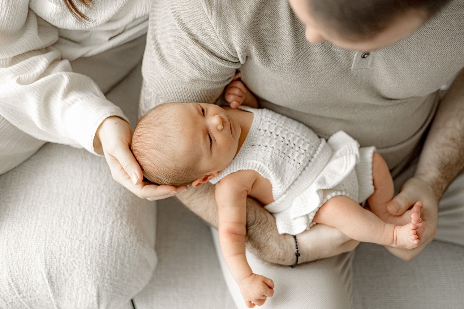 A newborn baby sleeping peacefully in a parent's arms, dressed in a white knit outfit.