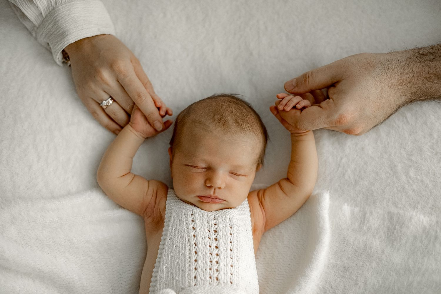 Newborn baby sleeping peacefully in white knit outfit while parents gently hold tiny hands on soft white blanket.