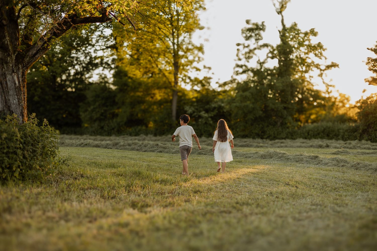 Two children run hand-in-hand through a lush green field bathed in golden sunset light.