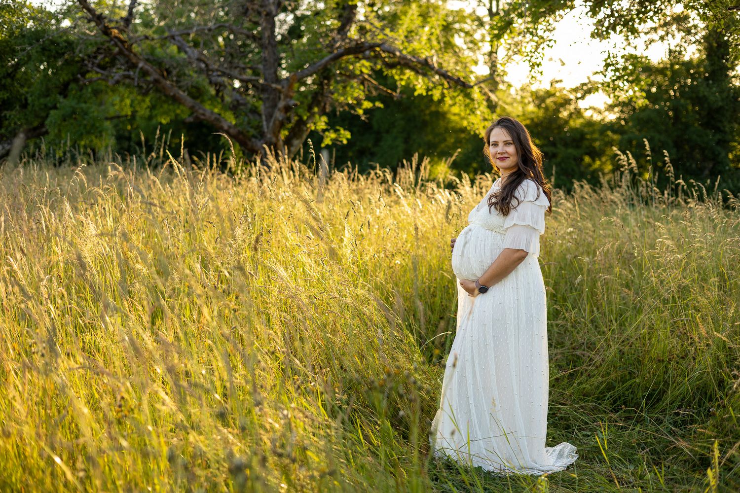 Pregnant woman in white dress standing in golden sunlit field with tall grass and trees during maternity photo session.