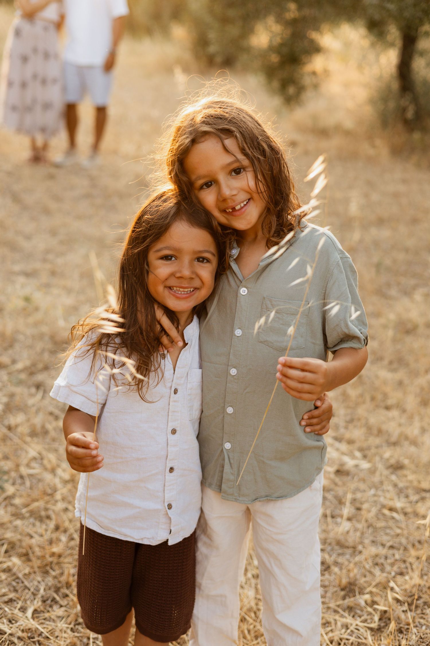 Two smiling children hugging outdoors in golden sunlight, wearing casual shirts in a dry grassy field.