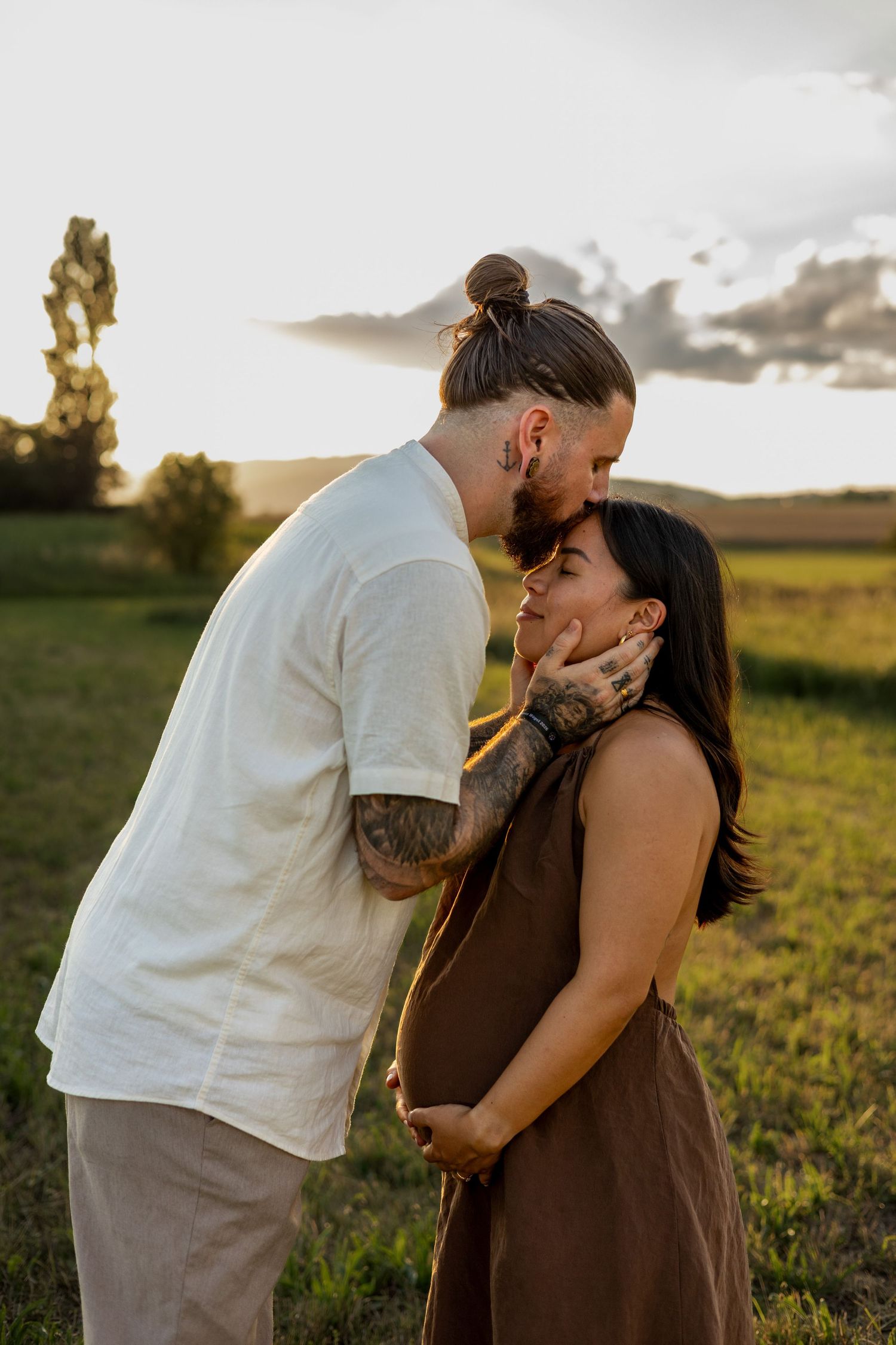 Couple sharing a tender moment at sunset in a green field, man gently holding pregnant woman's face.