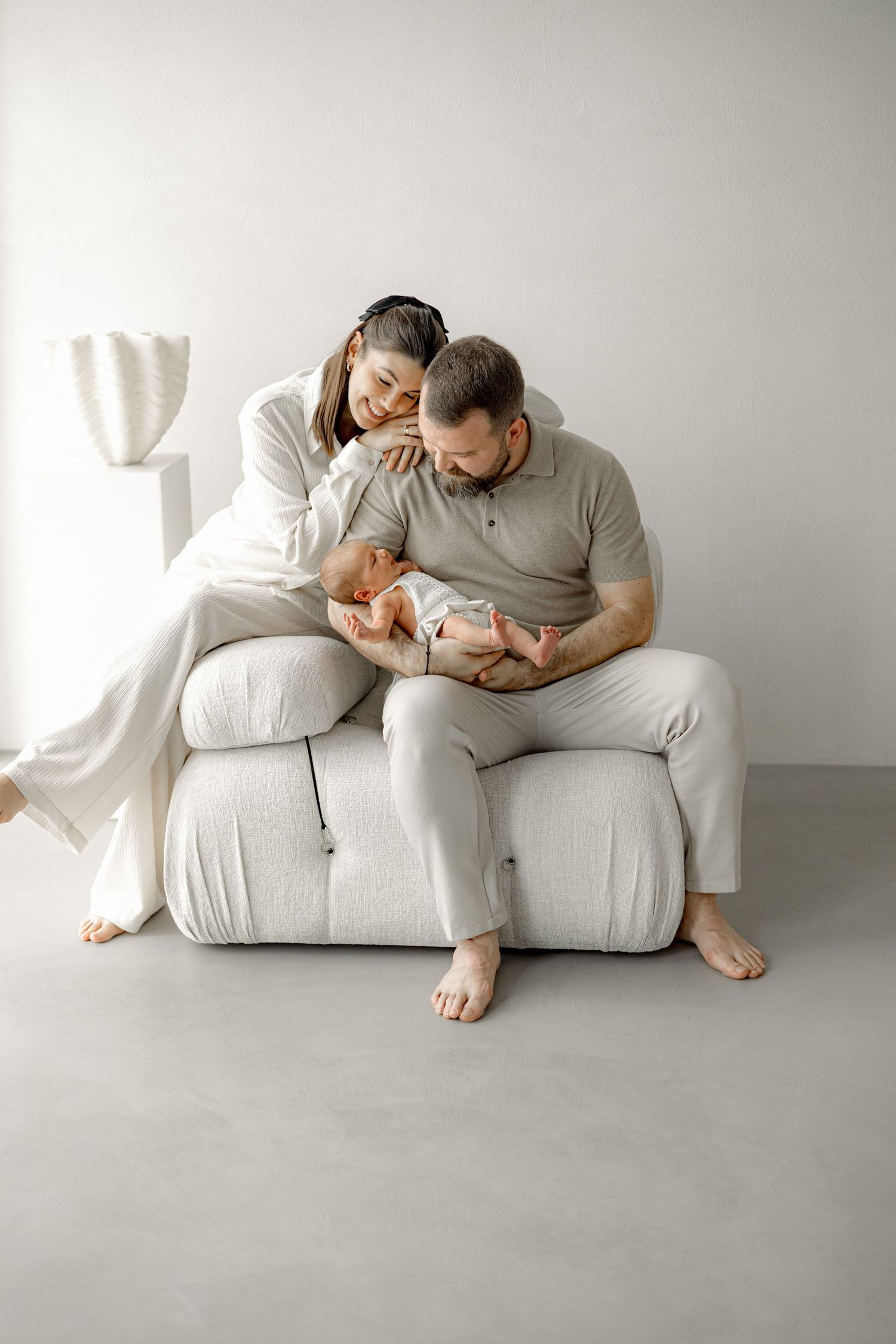 A couple in white outfits sits on a cream chair, lovingly gazing at their newborn baby in a bright studio.