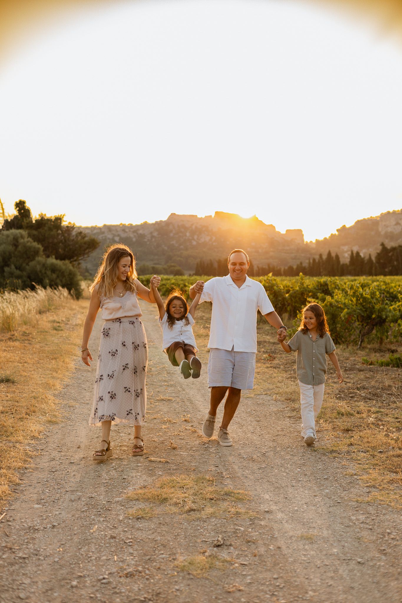 Family of four walking on a sunny dirt path through vineyards at golden hour, parents swinging child between them.