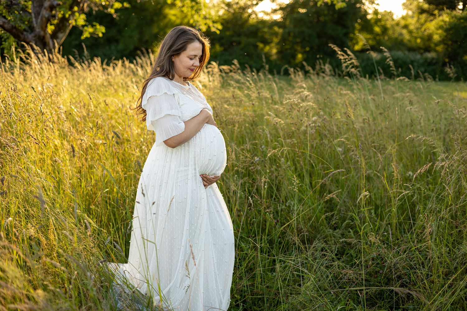 Pregnant woman in white dress cradling baby bump in golden sunlit meadow during maternity photo session.