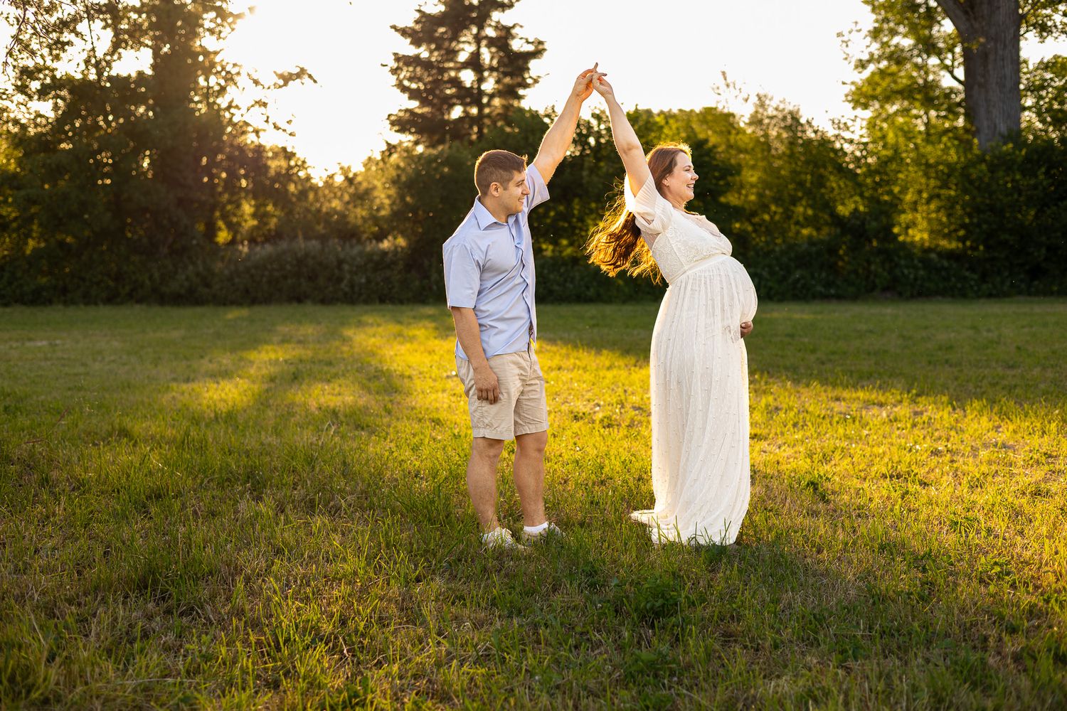 Pregnant woman in white dress dancing with partner in sunny green field during golden hour maternity photo session.