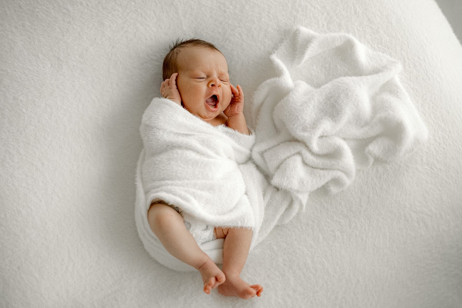 Newborn baby wrapped in white blanket yawning while lying on a soft white surface.