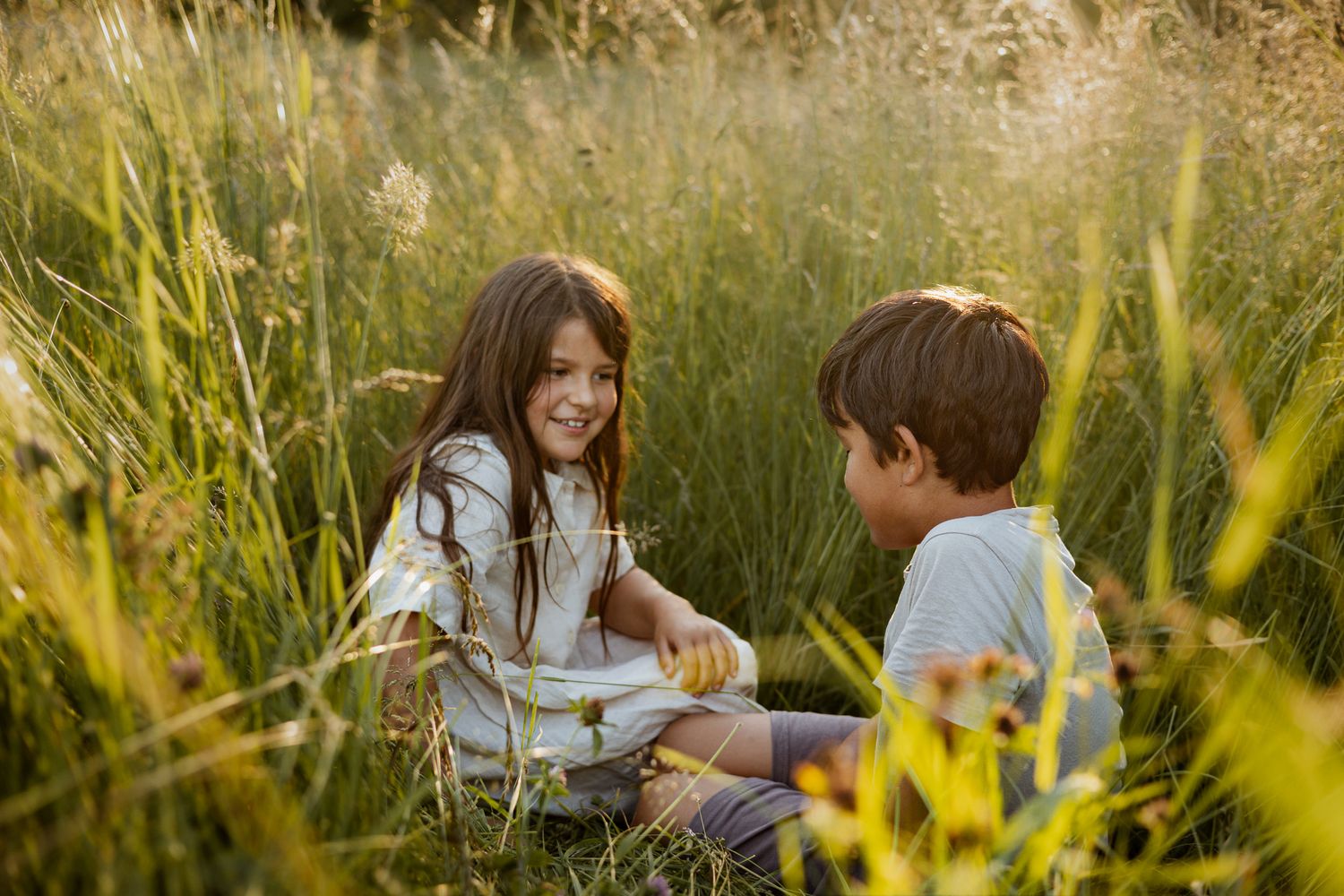 Two happy children sitting together in tall green grass field during golden hour sunlight.