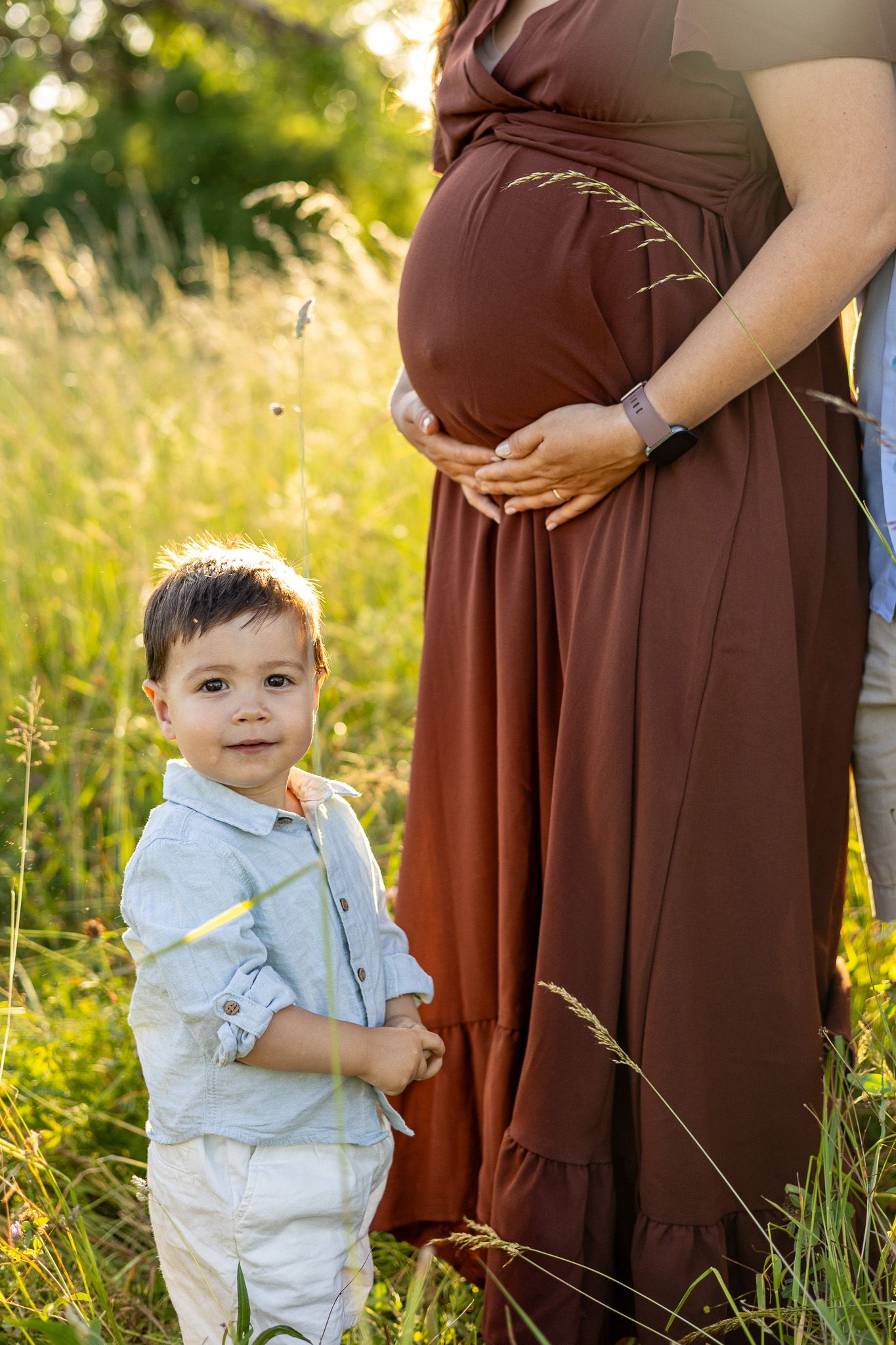 A toddler boy stands beside a pregnant woman in a brown dress in a golden sunlit meadow.