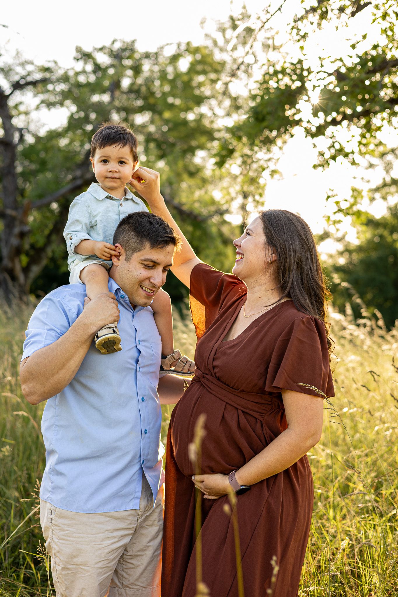 Pregnant woman in rust dress smiles at toddler on dad's shoulders in a sunlit green field.