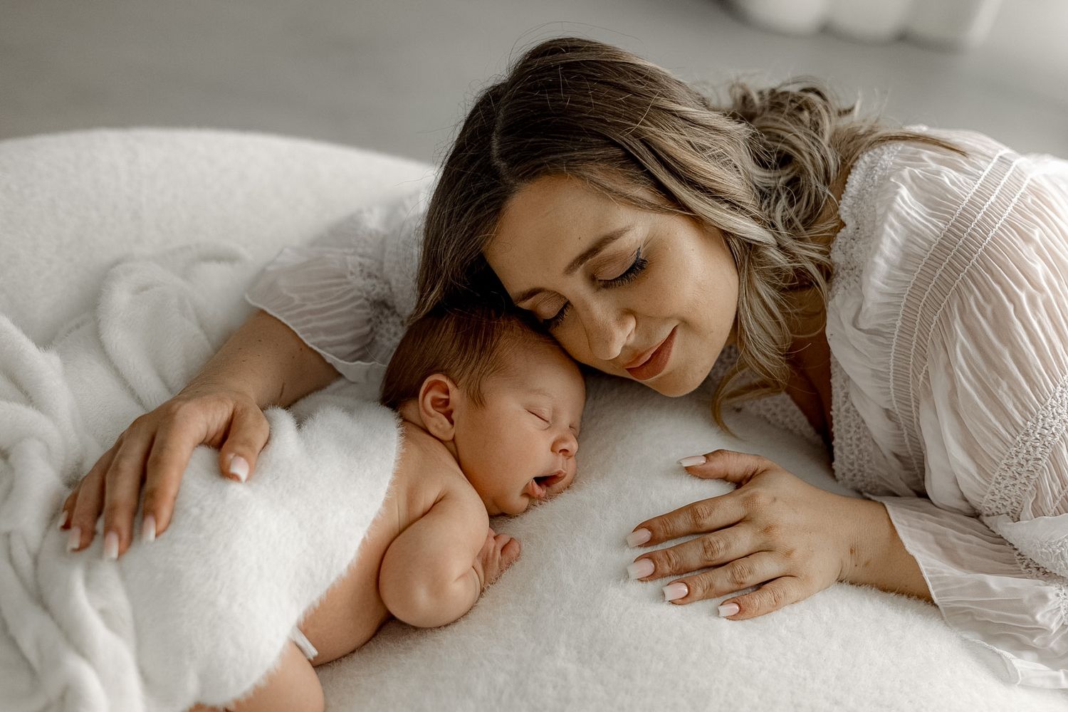 A mother tenderly rests her head beside her sleeping newborn baby on a soft white fluffy surface.
