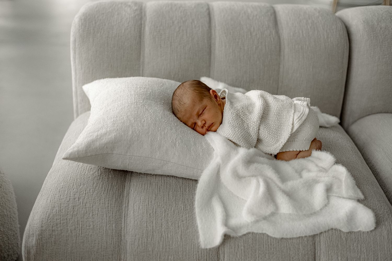 Newborn baby sleeping peacefully on a white sofa, wrapped in a soft white blanket with a pillow.