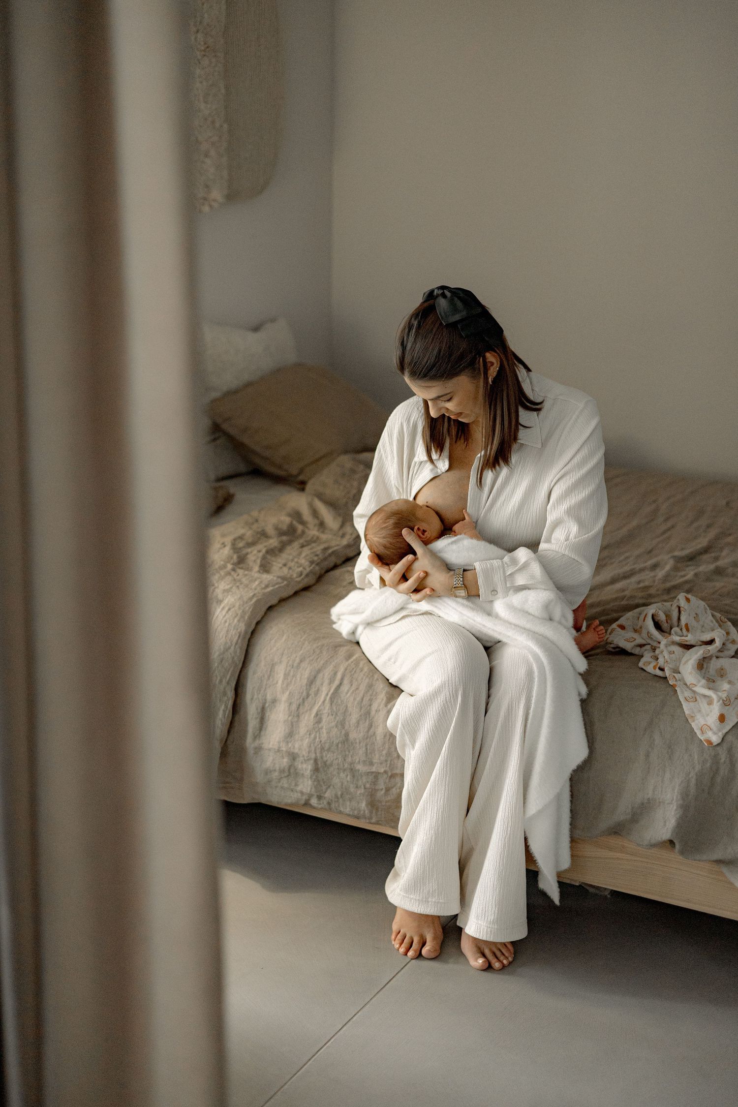 A mother in a white robe sits on a bed breastfeeding her newborn baby in a serene, neutral-toned bedroom.