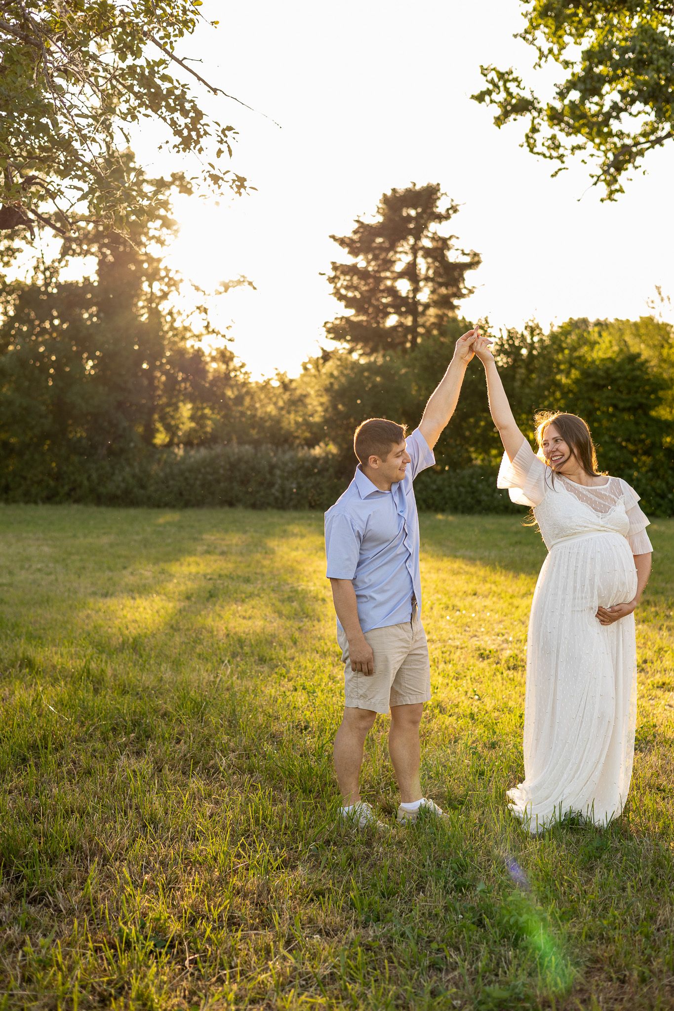 Pregnant woman in white dress dancing with partner in sunny park at golden hour.