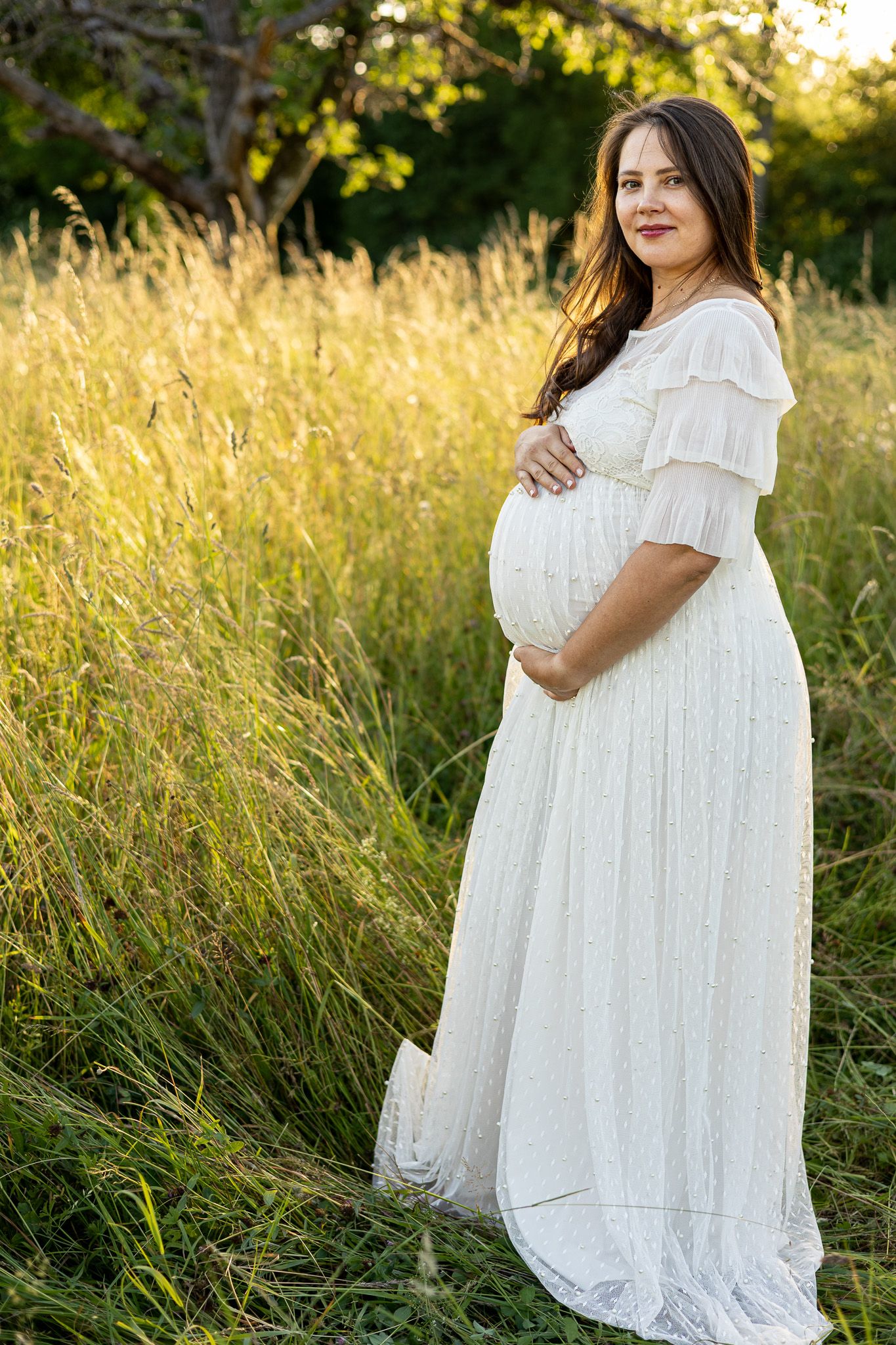 Pregnant woman in white lace dress holding baby bump in golden sunlit field.