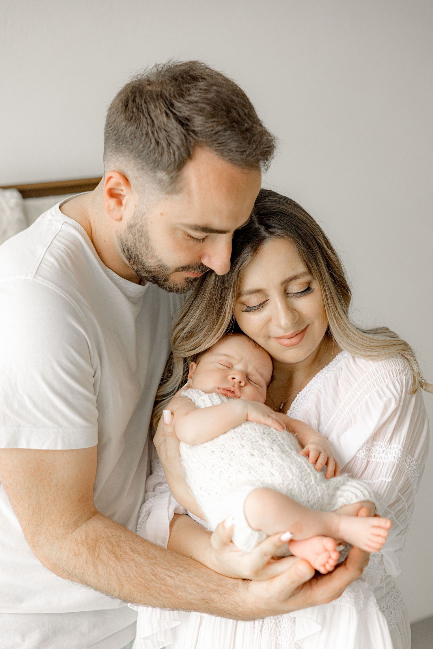 A couple in white clothing tenderly holds their sleeping newborn baby, eyes closed in a serene family portrait.