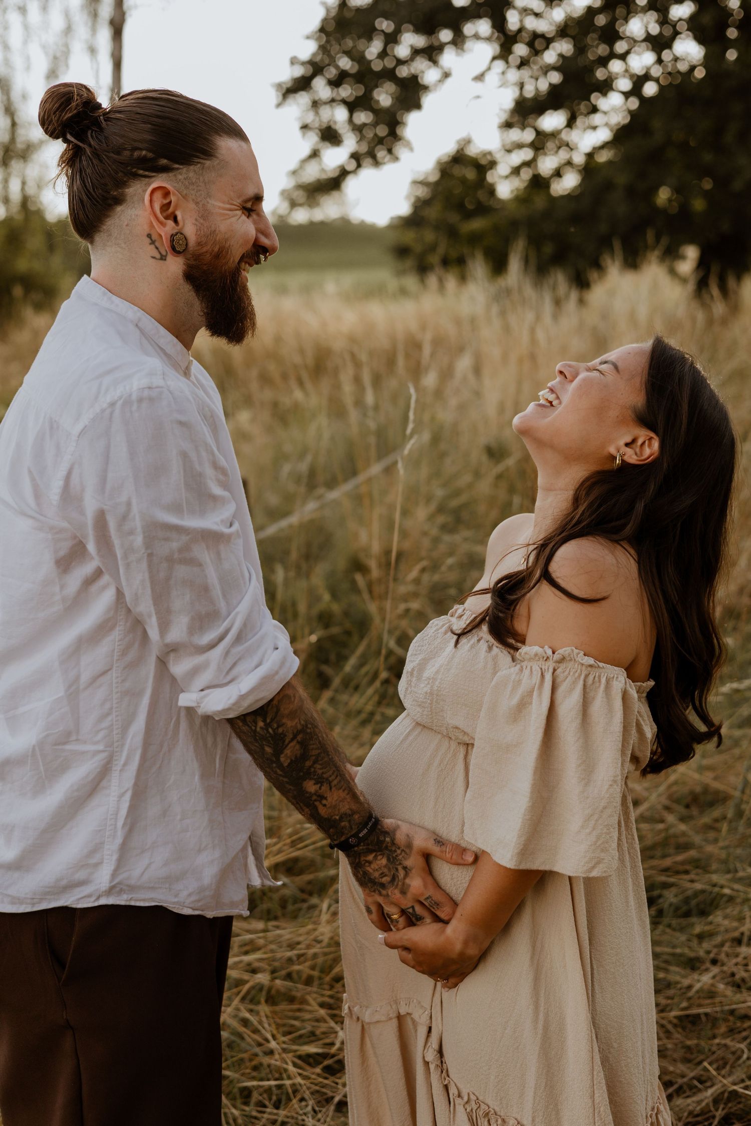 A laughing pregnant couple holds hands in a golden field, the man with tattoos and a bun, the woman in an off-shoulder dress.