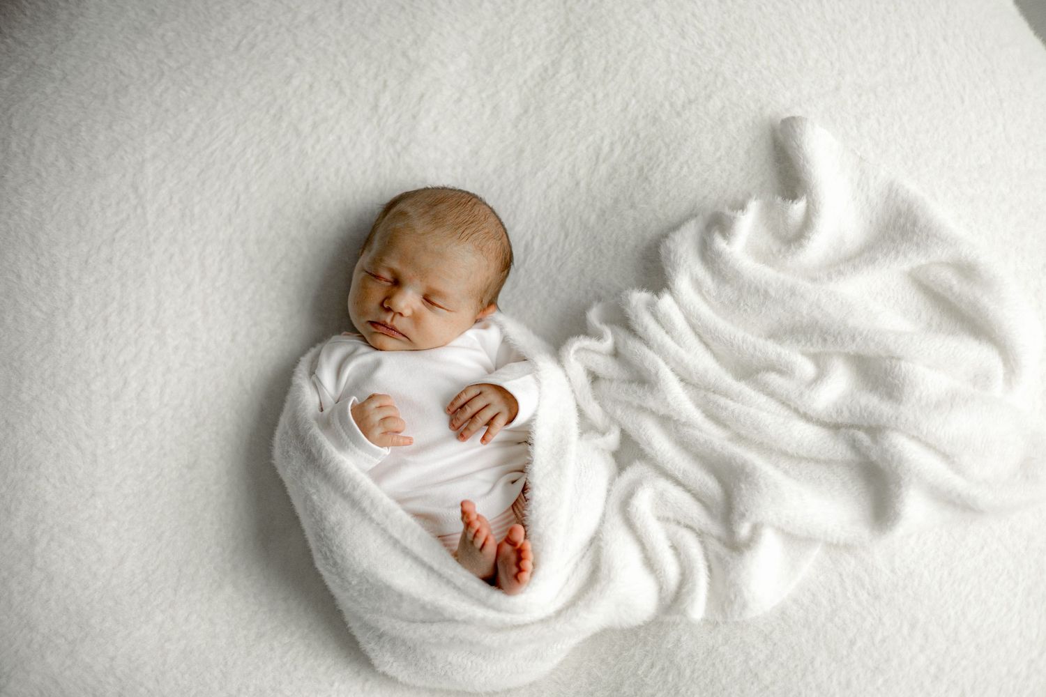 Newborn baby swaddled in white blanket sleeping peacefully on a soft white background.