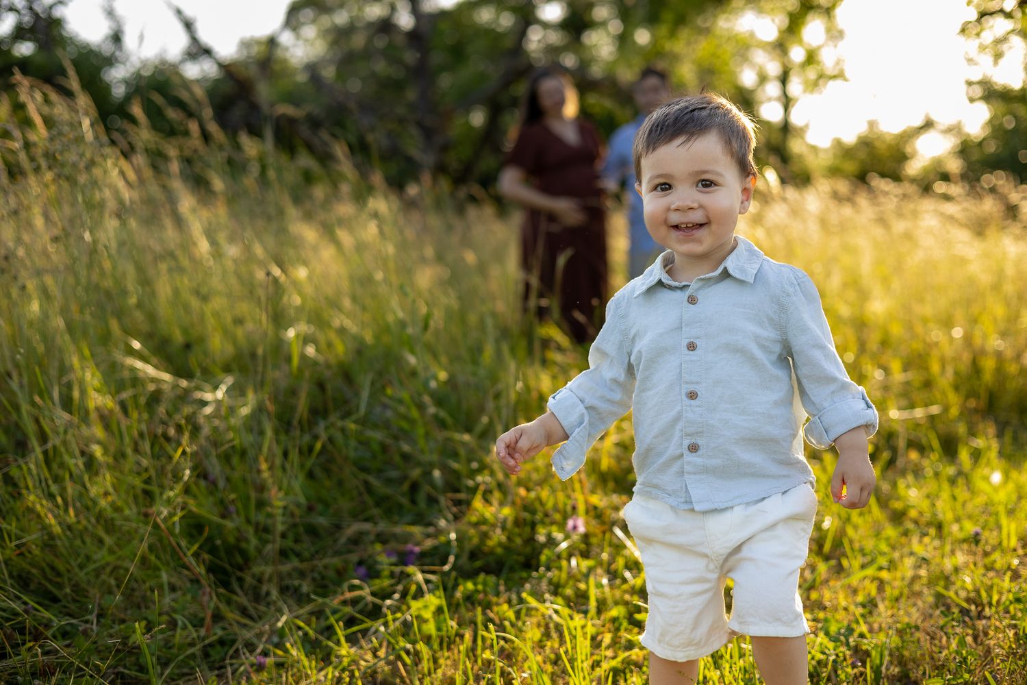 A happy toddler boy in a light blue shirt runs through a sunny green meadow with family in the background.