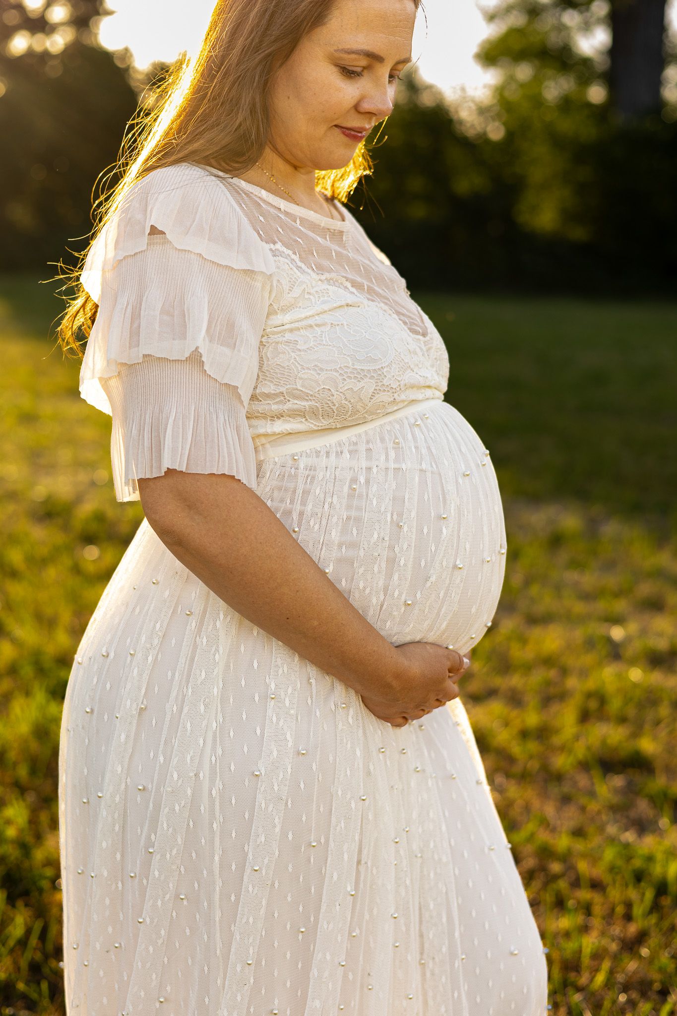 Pregnant woman in white lace dress cradling baby bump outdoors in golden sunset light.