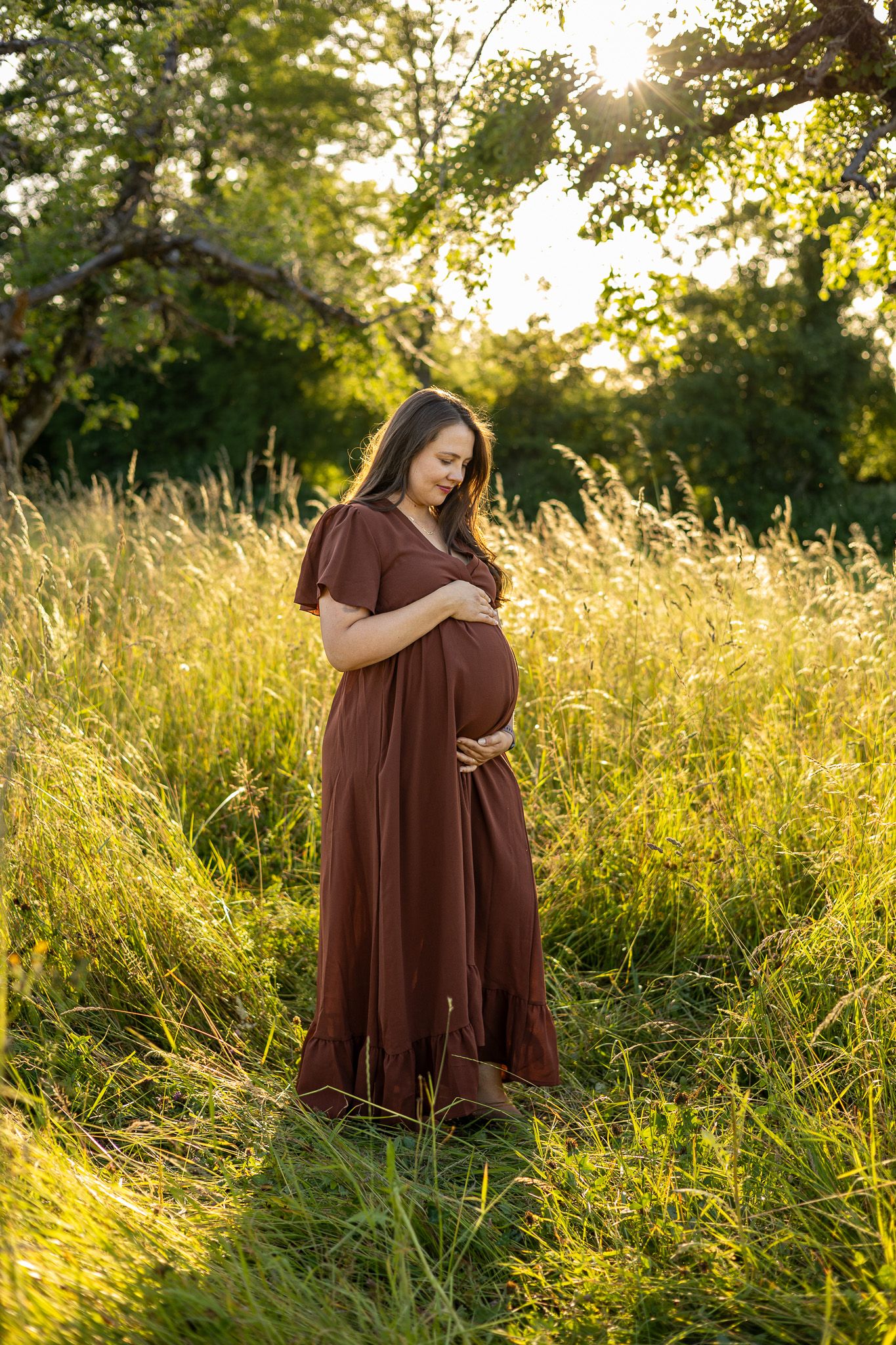 Pregnant woman in brown maxi dress standing in sunlit field, cradling baby bump during golden hour maternity photo session.