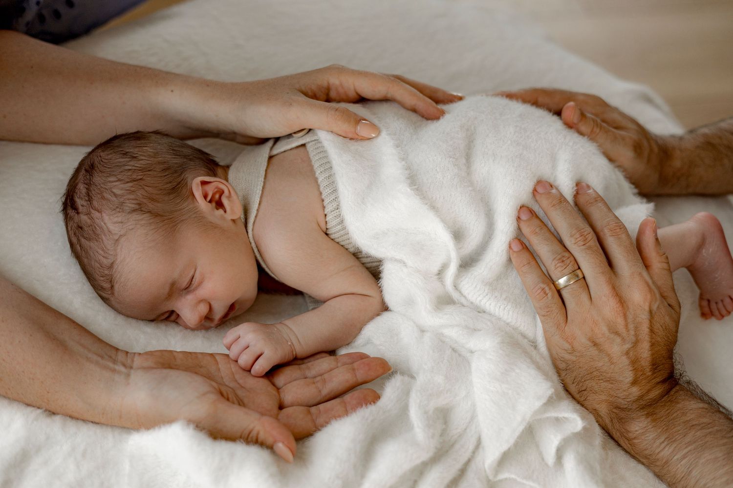 Newborn baby sleeping peacefully on white blanket while being gently held by two pairs of adult hands.