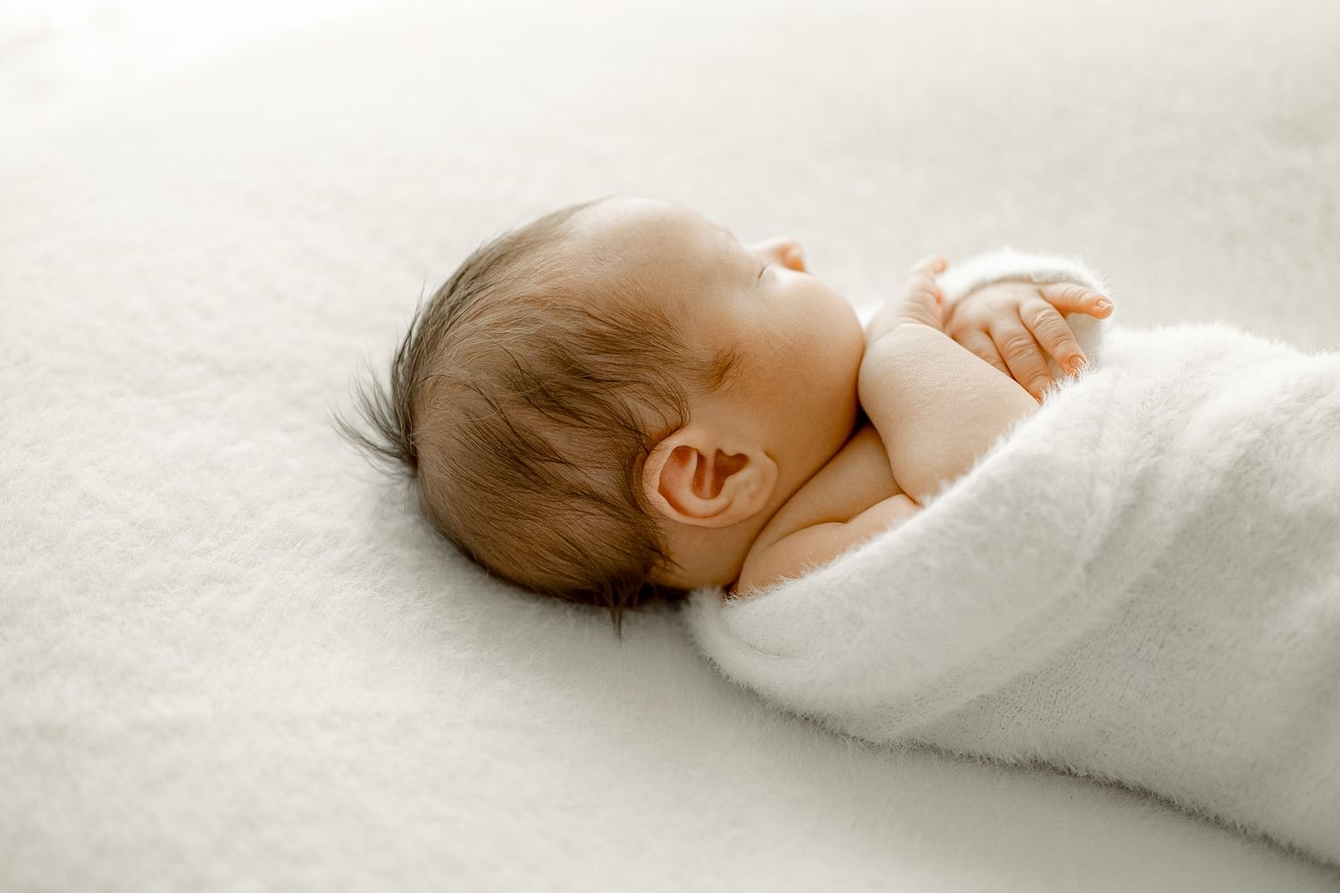 Peaceful newborn baby sleeping wrapped in a soft white blanket on a white surface.