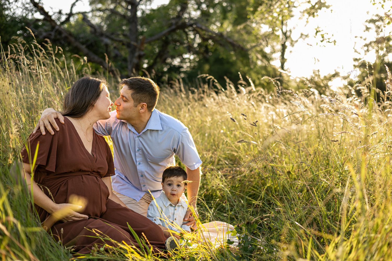 Pregnant woman and husband share a kiss while sitting with toddler son in golden sunlit meadow grass.