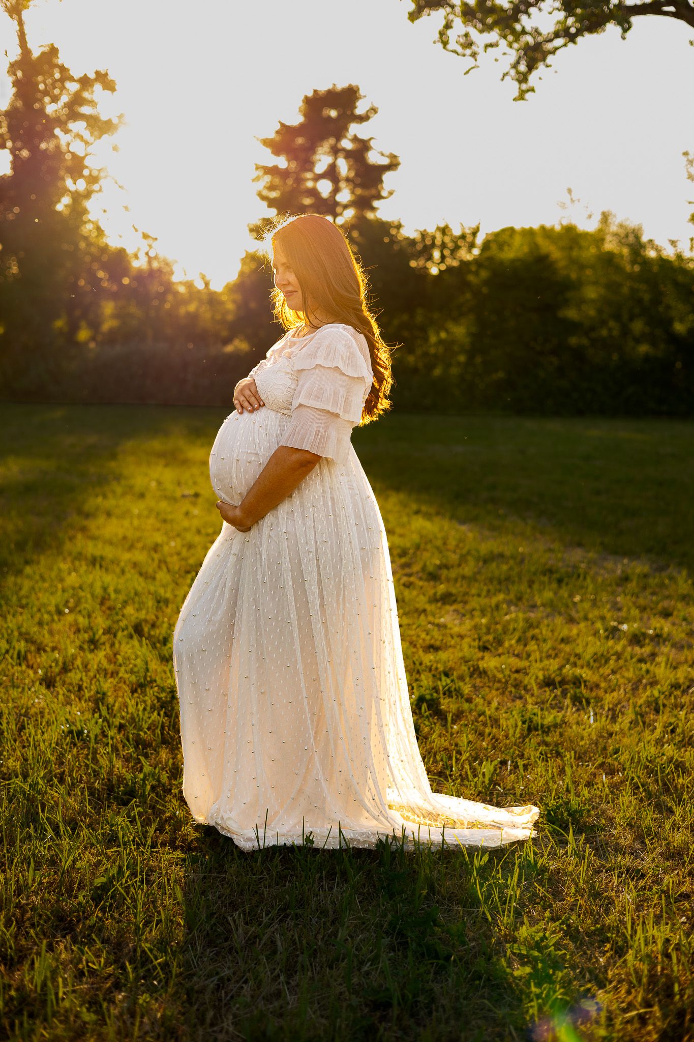 Pregnant woman in white lace dress cradling baby bump in sunlit green field at golden hour.