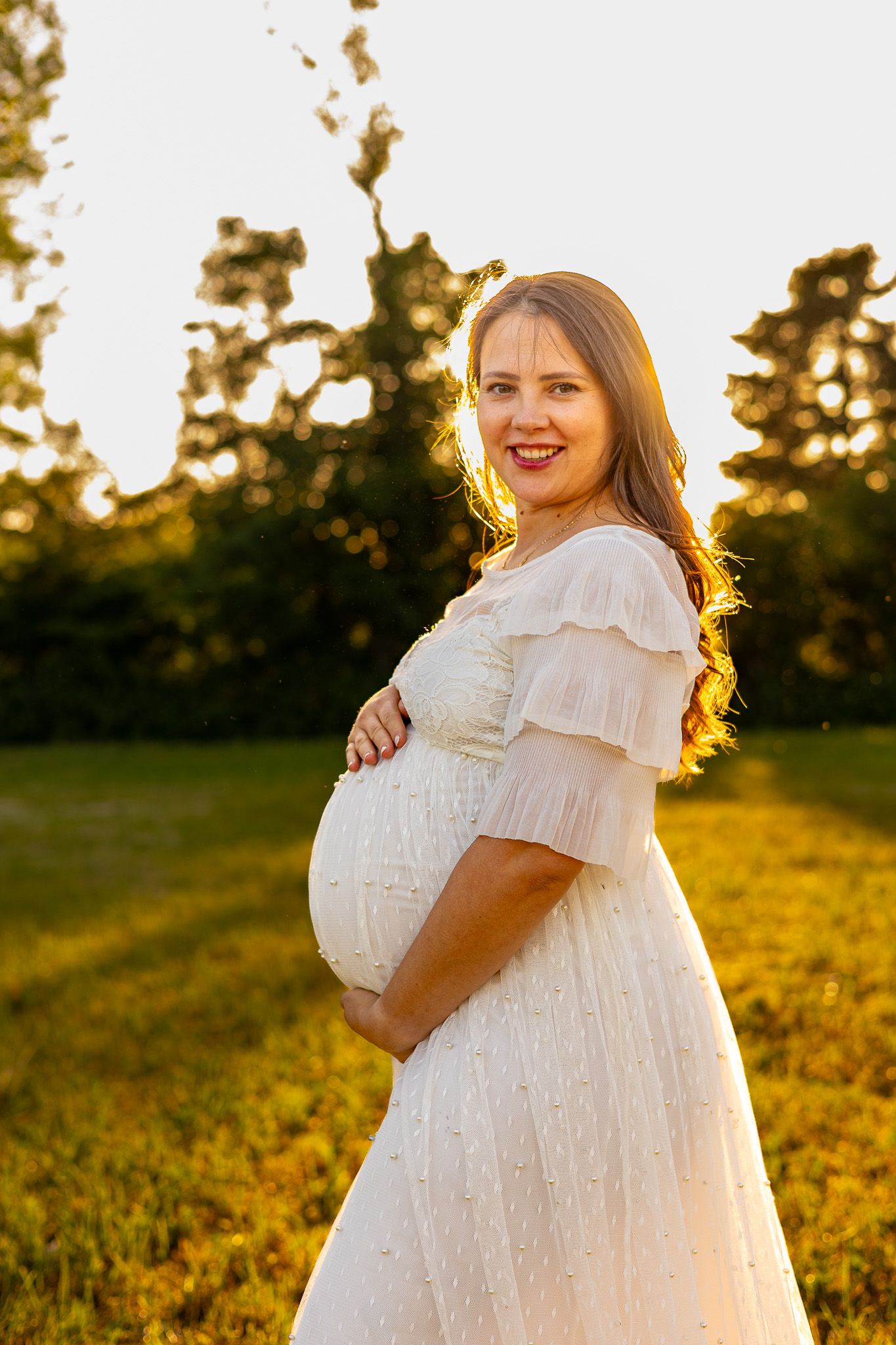 Pregnant woman in white dress holding baby bump outdoors at golden sunset hour.