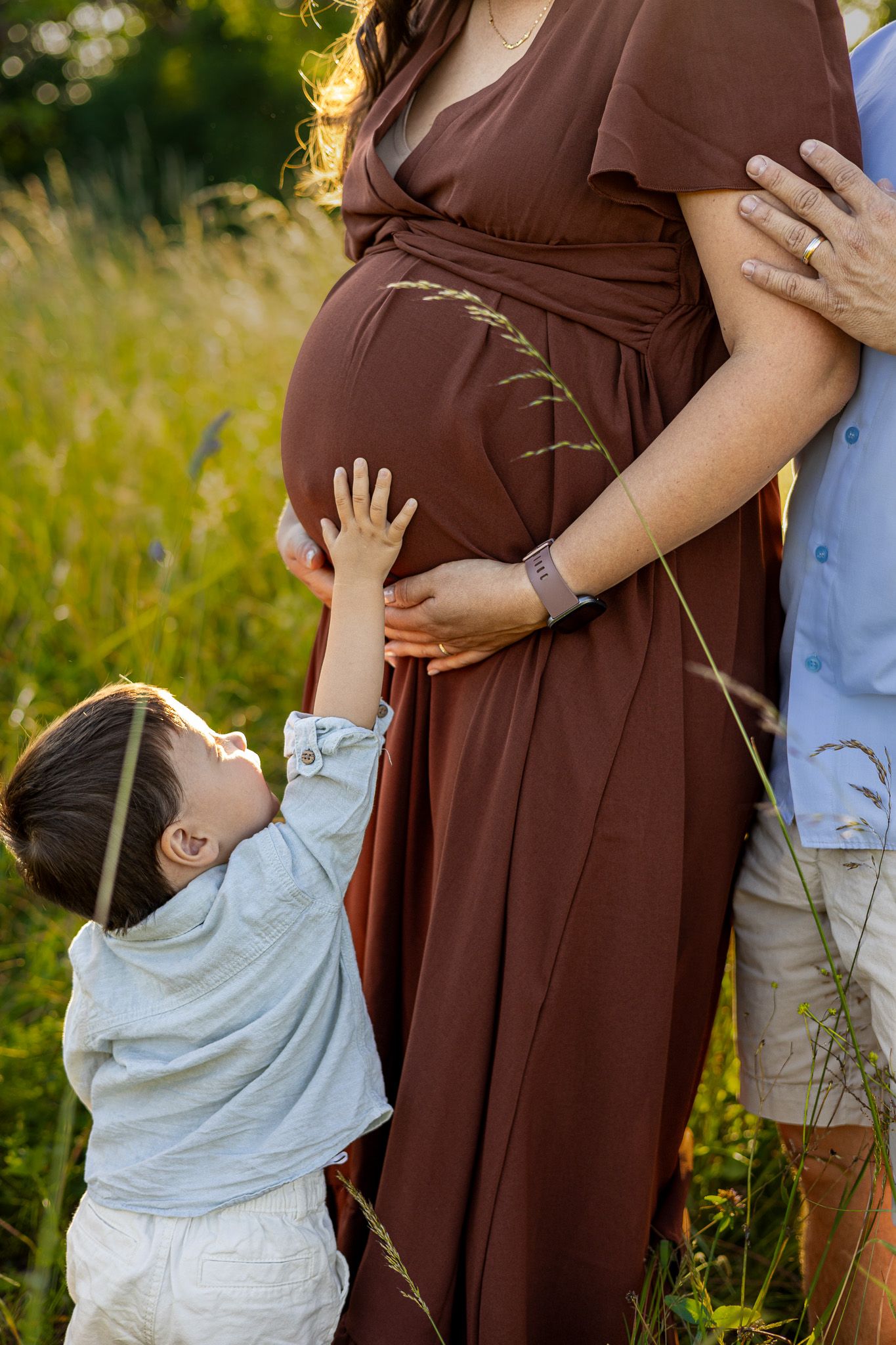 A young child touches a pregnant woman's belly in a sunlit field, as a man stands beside her.