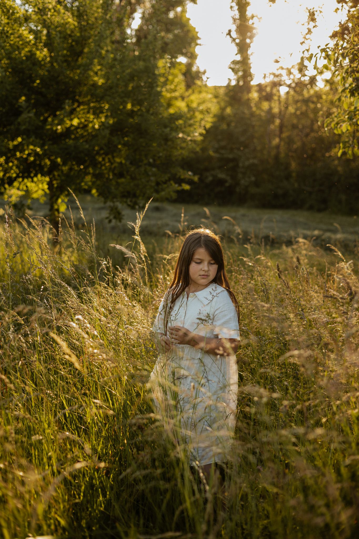 Young girl in white dress standing in tall golden grass field at sunset with green trees in background.