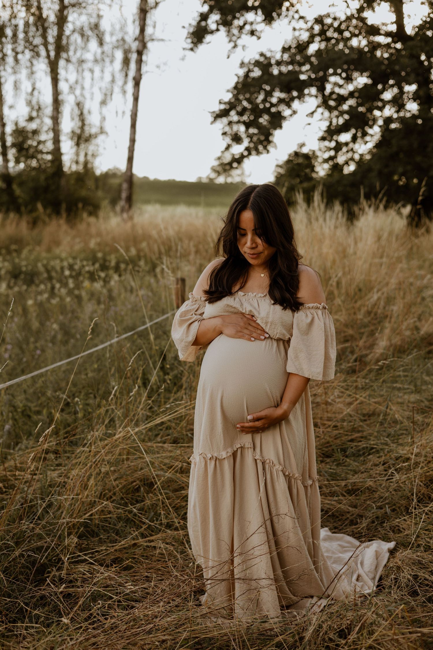 Pregnant woman in beige off-shoulder maxi dress cradling baby bump in golden hour field setting.