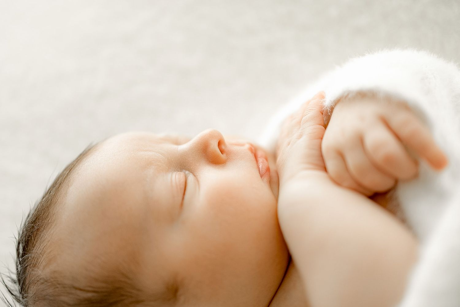 Peaceful newborn baby sleeping soundly, wrapped in a soft white blanket, with tiny hands near face.