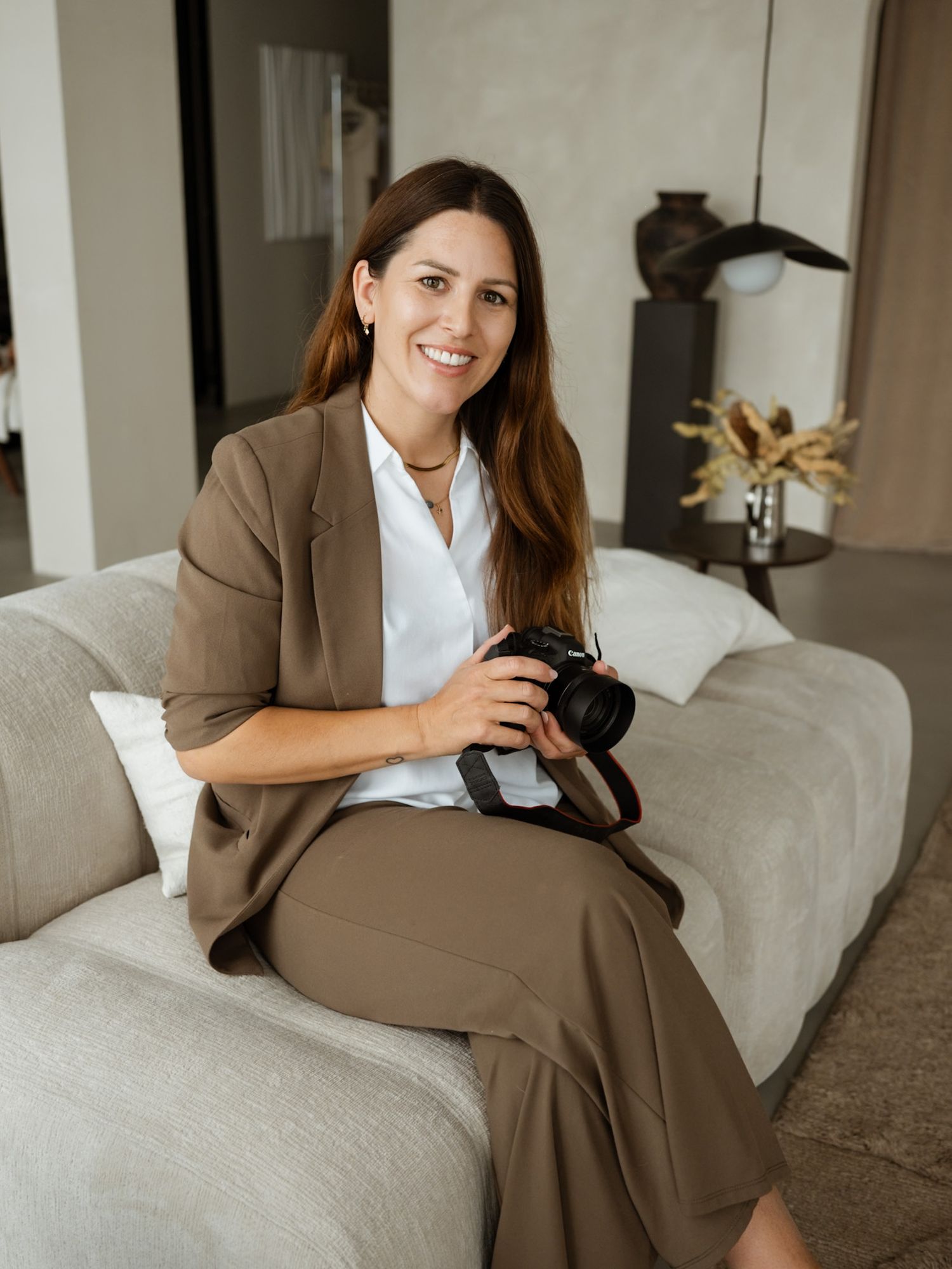 Woman in tan suit sitting on sofa holding a professional camera, smiling in a modern interior setting.