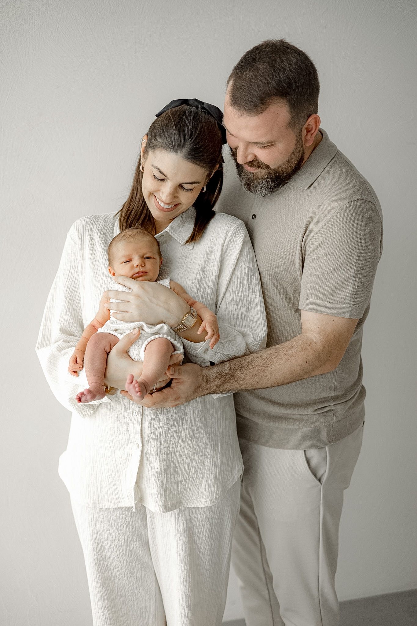Happy parents in neutral tones lovingly hold their newborn baby together against a white background.
