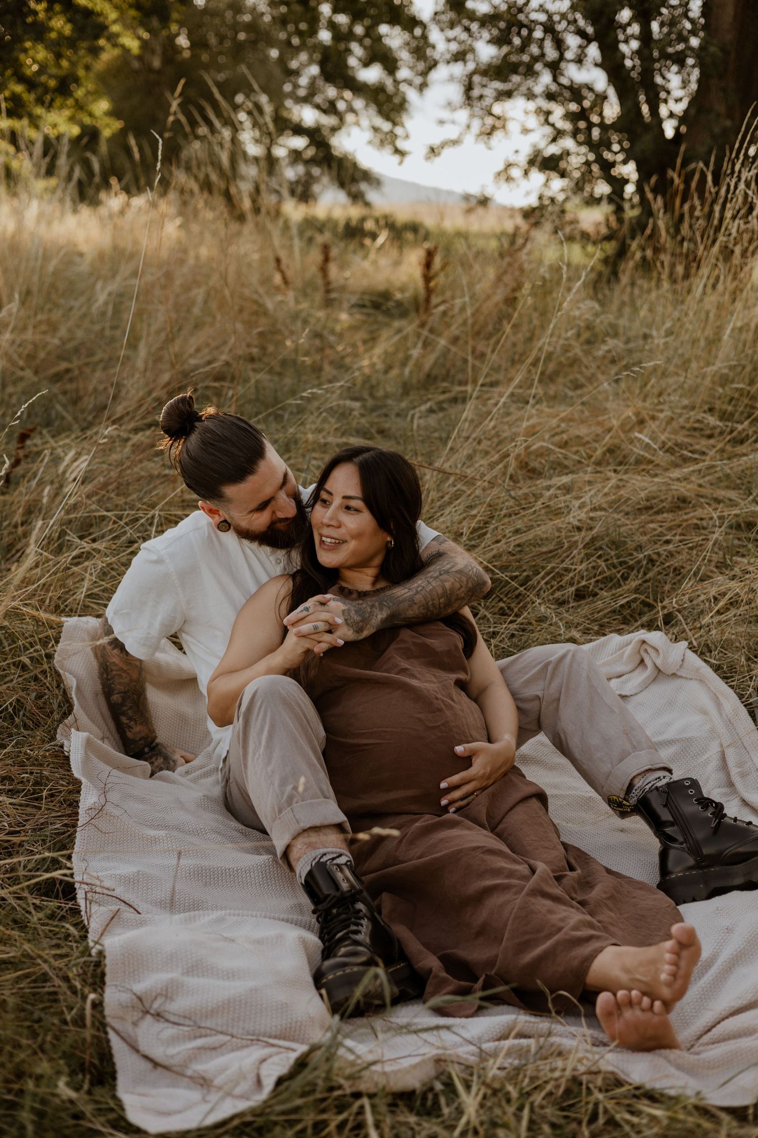 A couple relaxes on a blanket in a golden field, the woman smiling as her partner embraces her from behind.