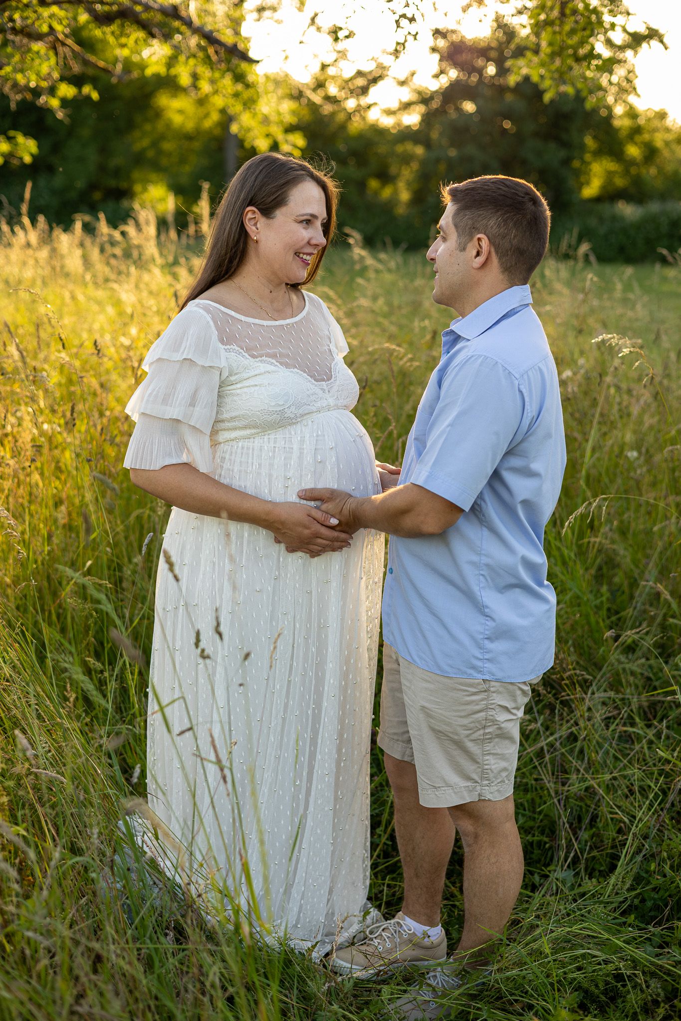 Pregnant woman in white dress and partner holding her belly in a sunlit field during golden hour maternity photoshoot.