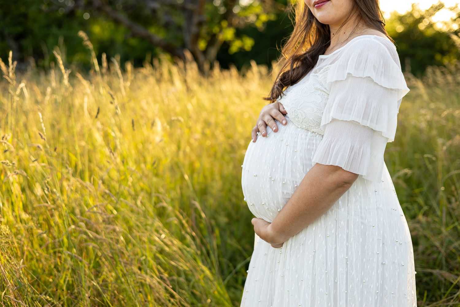 Pregnant woman in white dress cradling baby bump in golden sunlit field.