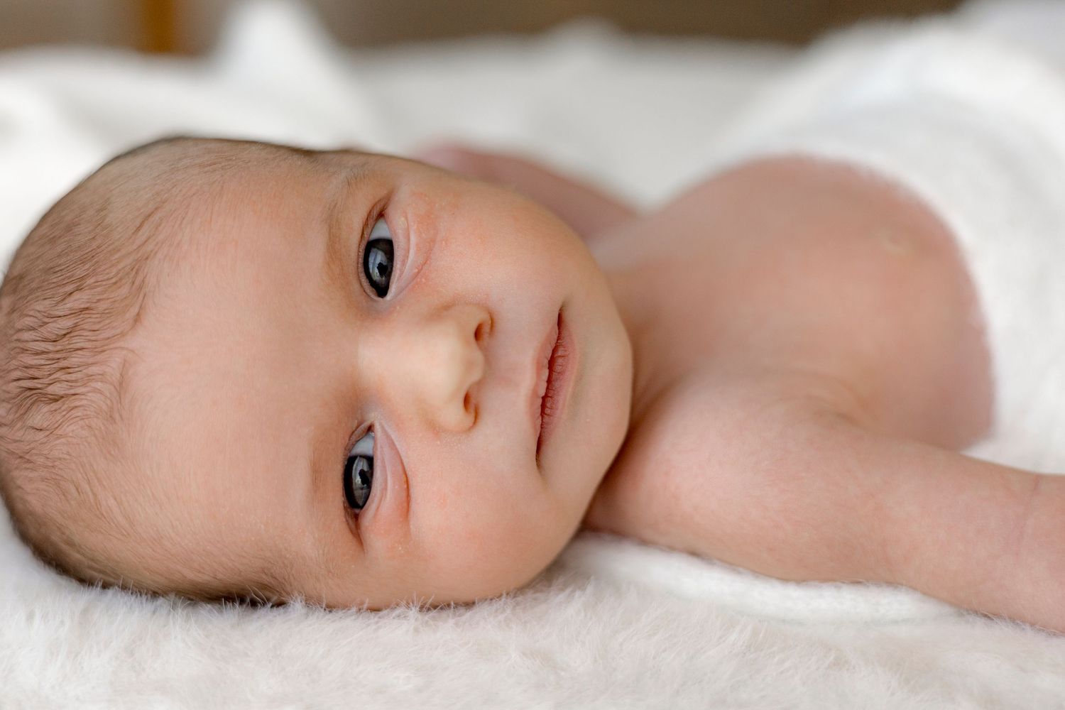 Newborn baby with blue eyes lying on a soft white blanket, looking peacefully to the side.