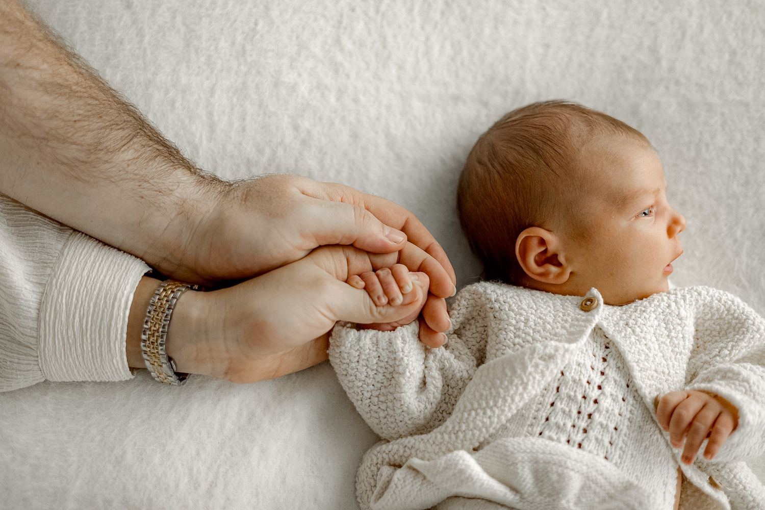 A newborn baby in a white knit outfit lies on a soft blanket while an adult gently holds their tiny hand.