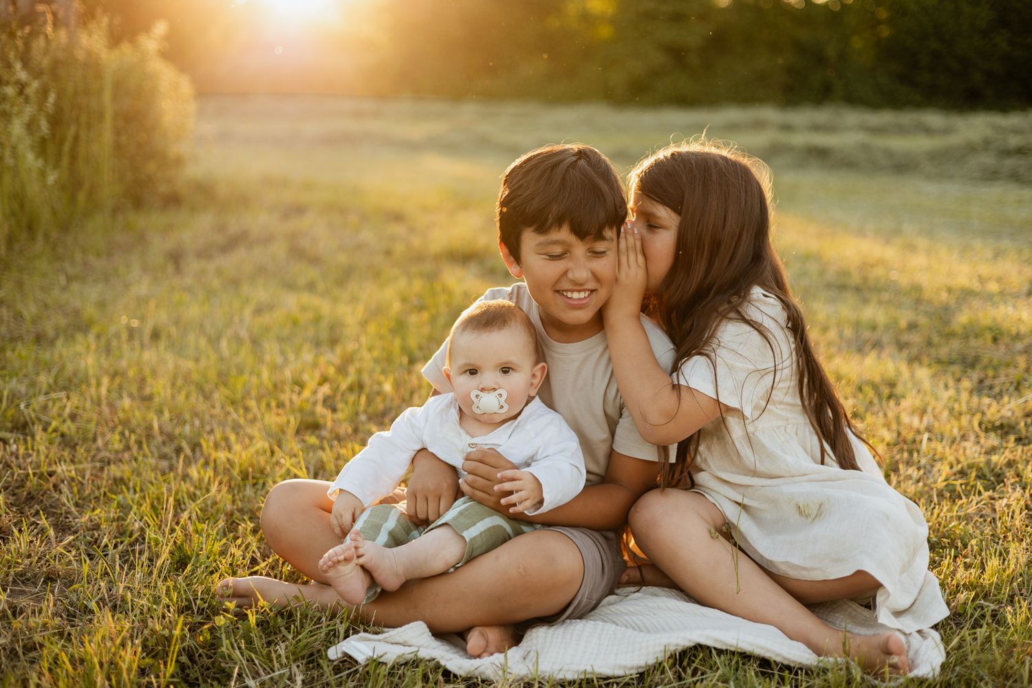 Three children sit on a blanket in a sunny field, with a girl whispering to a boy holding a baby with a pacifier.